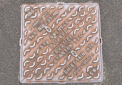 Square metal wastewater cover within the road, featuring a raised chevron pattern and the words SW. 