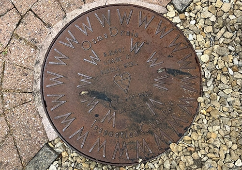 A metal circular wastewater cover, amongst the pavement. The metal cover has a raised pattern with the letter W around the outside and centre.