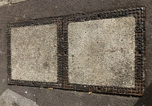 A metal rectangular telecoms cover, split into two squares with a metal frame and concreate centre. 