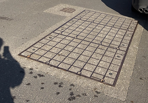 A large rectangular metal gattic cover, with a grid patter amongst the road surface. 