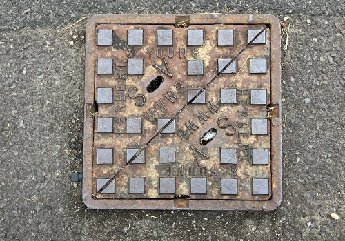 Square metal sluice valve cover set into a road surface, with raised square detailing and the letters 'SV' embossed on the cover.